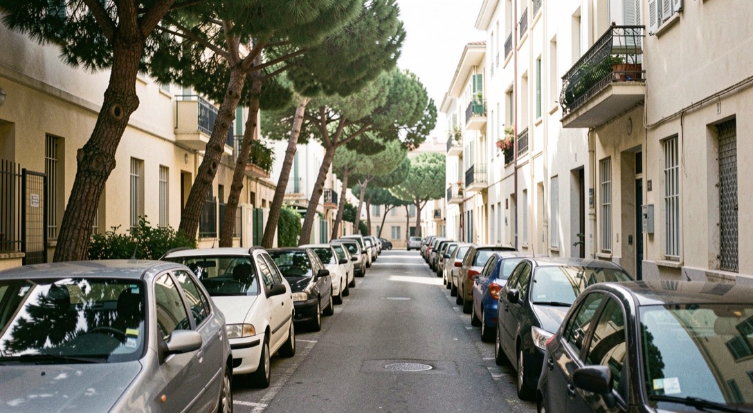 A narrow sloping residential street in an eastern Marseille neighborhood, cars parked tightly on one side, plane trees and pale apartment blocks, mid-morning